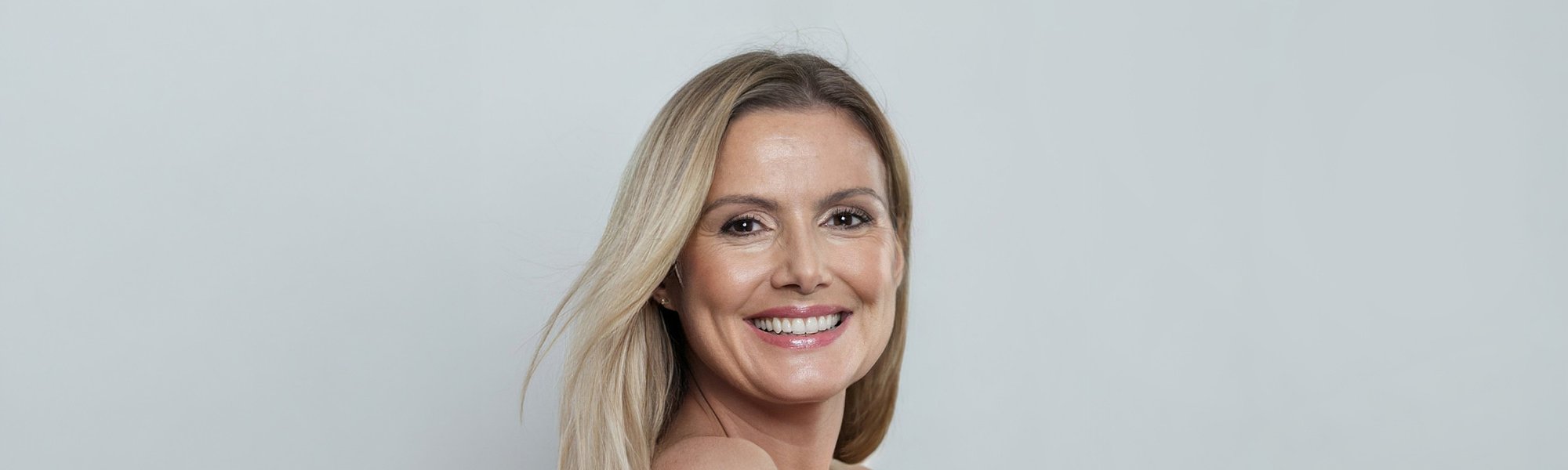 Portret uśmiechniętej kobty w średnim wieku. Ma długie, blond włosy i lekki makijaż na twarzy