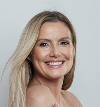 Portret uśmiechniętej kobty w średnim wieku. Ma długie, blond włosy i lekki makijaż na twarzy